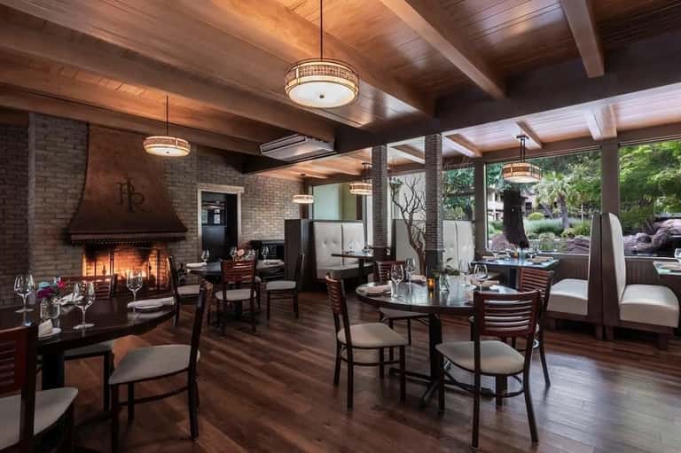 Upscale restaurant interior with exposed brick fireplace, wooden beams, dark furniture, and glass doors