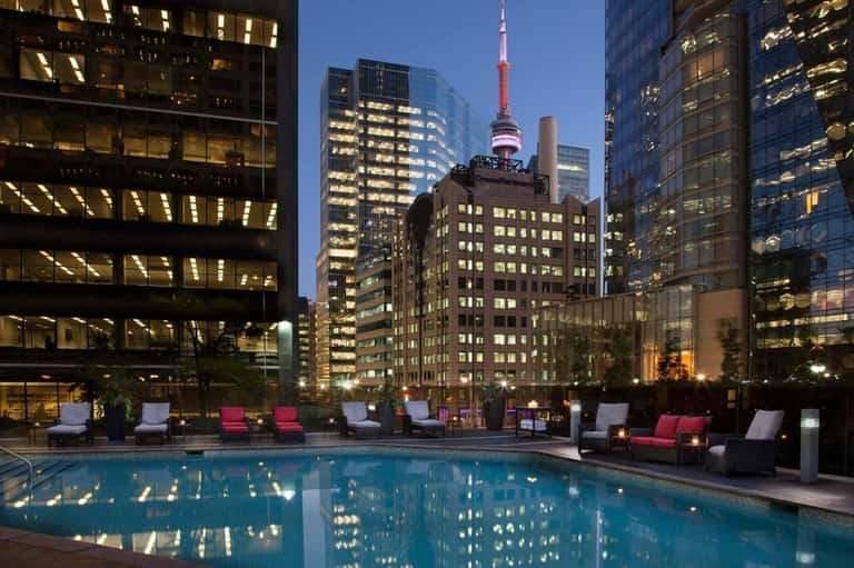 Urban rooftop pool at night reflecting city skyline, CN Tower visible, illuminated high-rise buildings surrounding deck