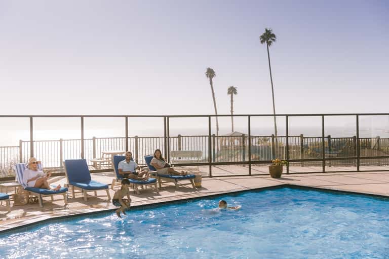 Rooftop pool deck with lounge chairs, palm trees, metal railing, and mountain backdrop at sunrise