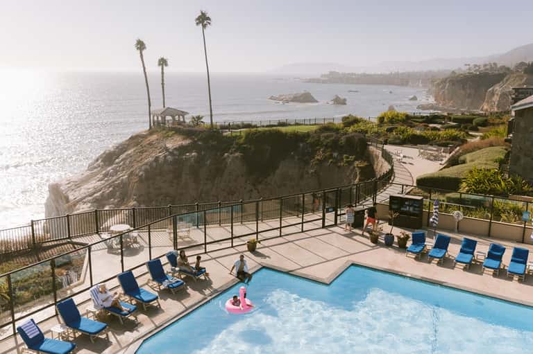 Cliffside pool overlooking ocean with blue lounge chairs, palm trees, and dramatic coastal landscape