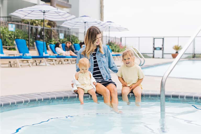 Woman and two toddlers at edge of shallow pool with umbrellas and lounge chairs visible in sunny resort setting