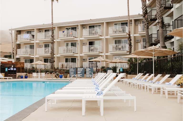 Three-story beachfront hotel with cream-colored balconies, palm trees, and white lounge chairs by pool