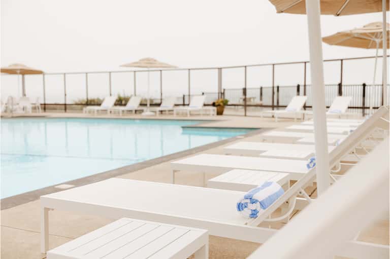 Resort pool deck with white loungers, tan umbrellas, glass railings, and ocean horizon in background