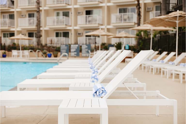 Resort poolside with white lounge chairs, umbrellas, and beige buildings