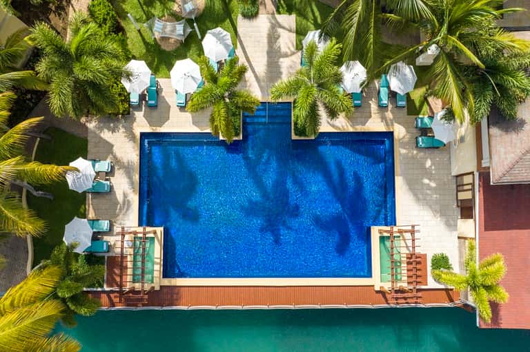 Aerial view of rectangular blue resort pool surrounded by white umbrellas and palm trees