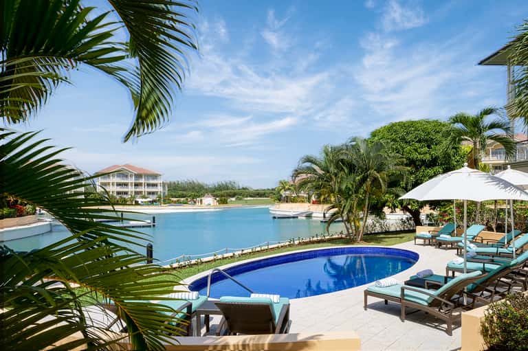 Resort pool area surrounded by palm trees, loungers, white umbrella, and lagoon with buildings