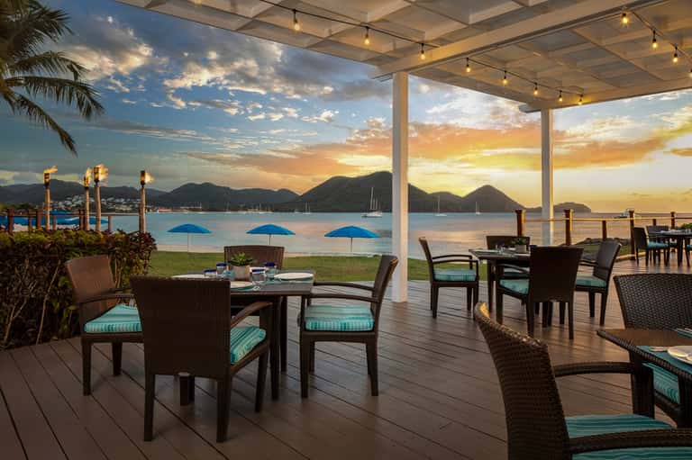 Beachfront dining area at sunset with mountain views, blue umbrellas, and wooden deck seating