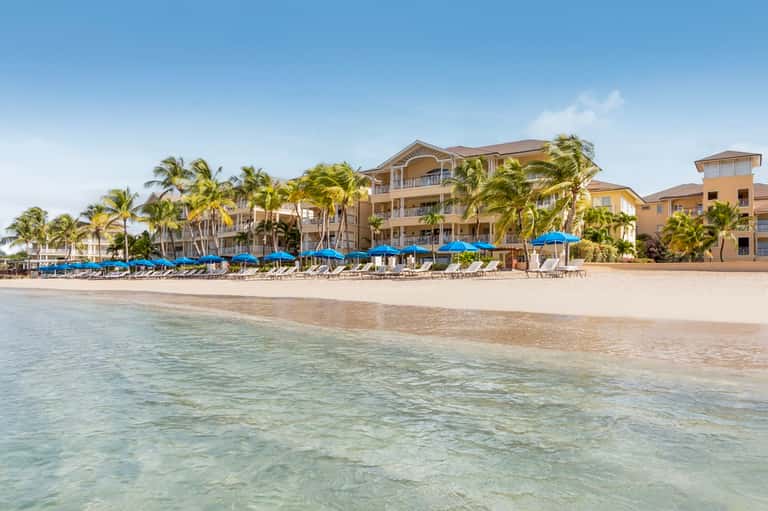 Beachfront resort with tan-colored buildings, palm trees, blue umbrellas, and sandy beach with shallow ocean water