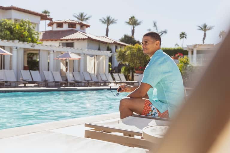 Guest relaxing by resort pool with chair lounges and Mediterranean-style white buildings with palm trees