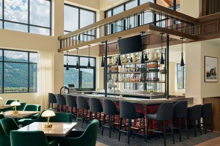 Modern lodge interior with floor-to-ceiling windows, mountain views, green armchairs, and elevated bar