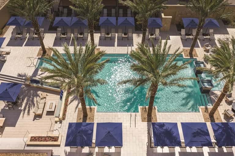 Aerial view of resort pool surrounded by palm trees, lounge chairs, and blue umbrellas