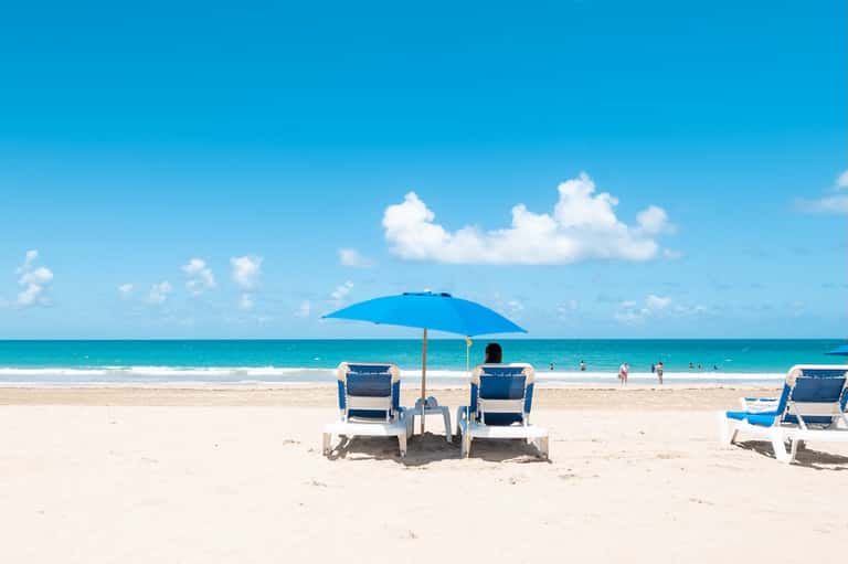 Pristine sandy beach with turquoise ocean, beach chairs under blue umbrella, and swimmers in the water