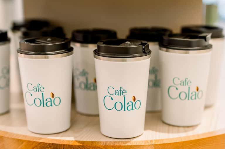 Row of white Café Colao branded to-go coffee cups with black lids on wooden surface