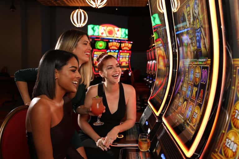 Three women playing slot machines in a brightly lit casino with colorful neon lights