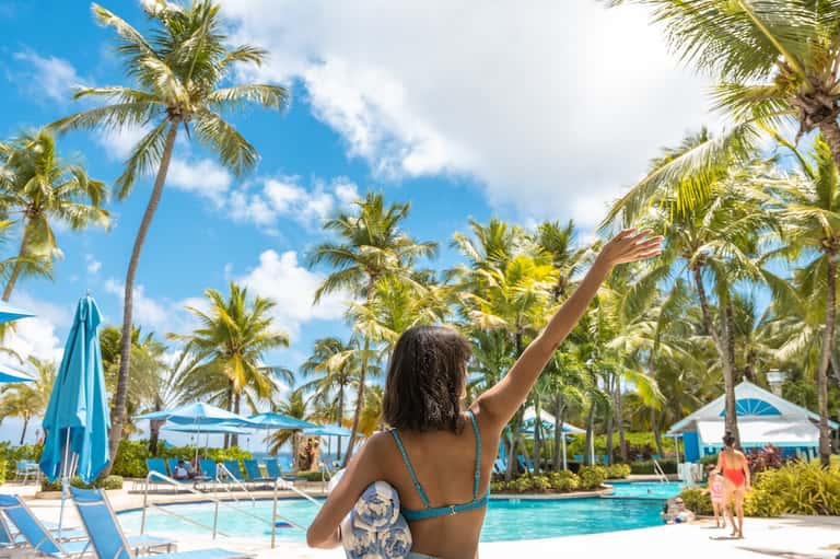 Woman in swimwear at tropical resort pool surrounded by palm trees and blue umbrellas