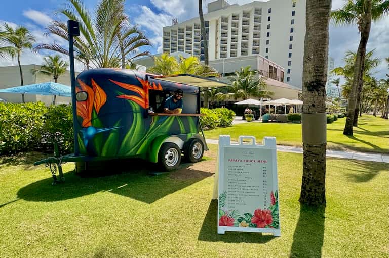 Colorful papaya food truck on resort lawn with palm trees and multi-story hotel building in background