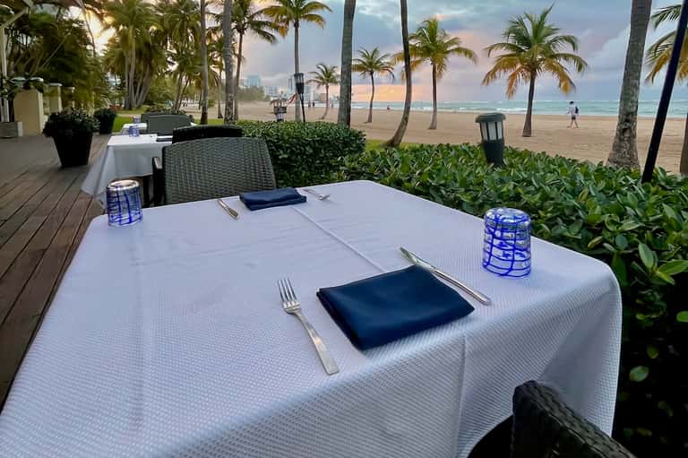 Beachfront dining table set for dinner with ocean, palm trees, and sandy beach visible in background