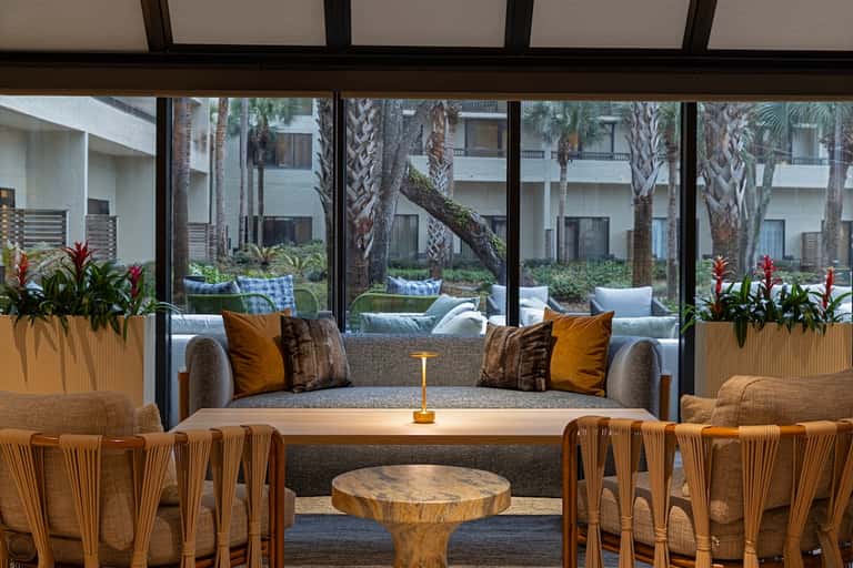 Resort lounge with gray sofa, woven chairs, tropical plants, and large windows overlooking palm-tree courtyard