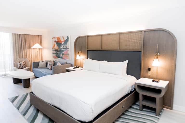 Contemporary hotel bedroom with wooden headboard, white bedding, and lounge seating area