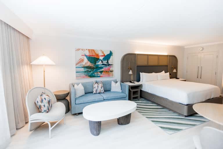 Modern hotel suite with blue sofa, two queen beds, ocean artwork, and balcony access