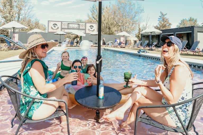 Family and friends enjoying poolside gathering with drinks at table near resort pool and buildings