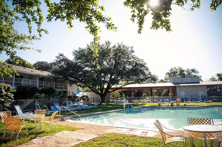 Resort pool area with loungers, shade structures, mature oak trees, and stone buildings on sunny day