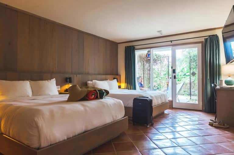 Hotel bedroom with two beds, wood paneling, glass patio doors, tropical garden view, and modern amenities