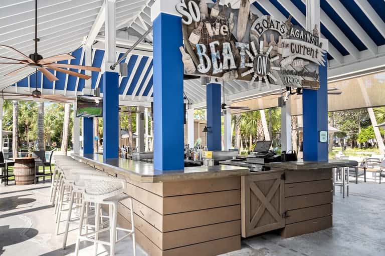 Beachfront tiki bar with blue columns, striped awning, white bar seating, nautical decor, and tropical surroundings
