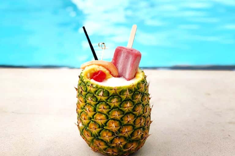 Tropical pineapple drink with pink popsicle on beach sand with ocean backdrop