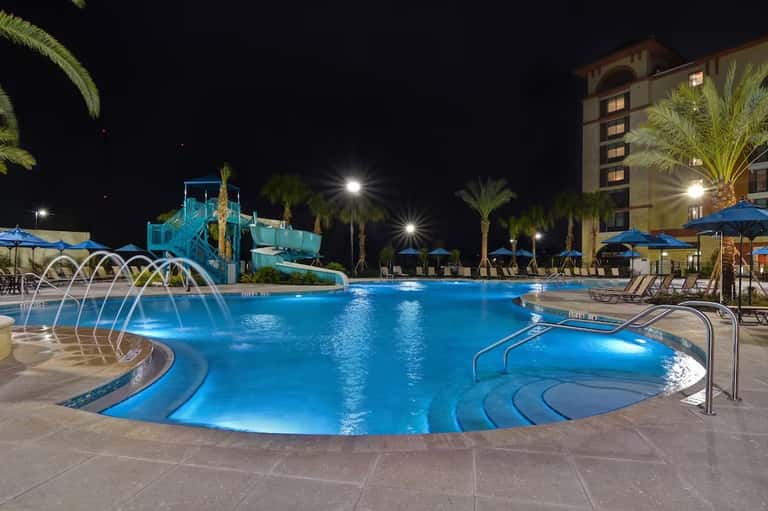 Illuminated resort pool at night with water slides, palm trees, and hotel building