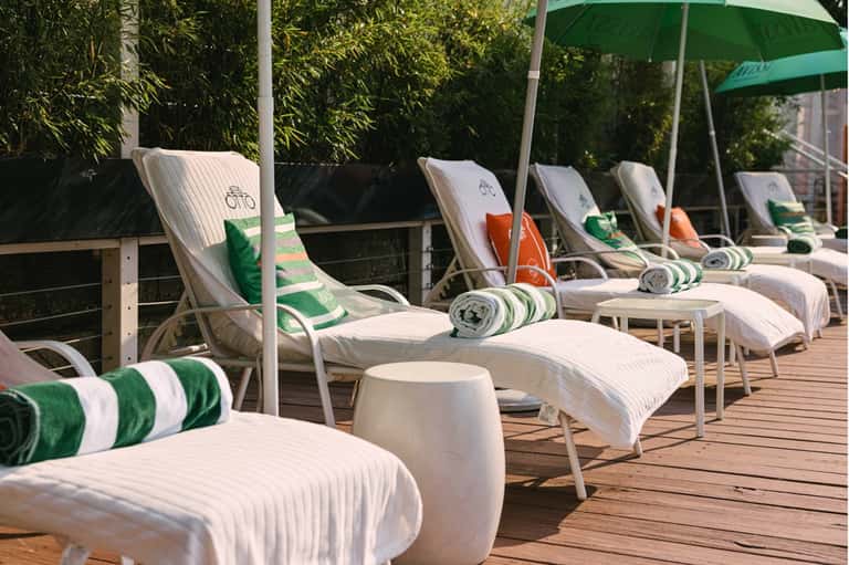 Poolside deck with white striped lounge chairs, green umbrellas, rolled towels, and bamboo privacy screen