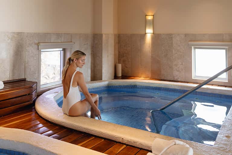 Woman in white swimsuit relaxing at indoor spa pool with natural light and tile accents