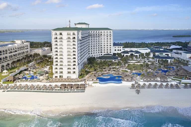 Waterfront resort with white main tower building, multiple pools, beach, and blue water views