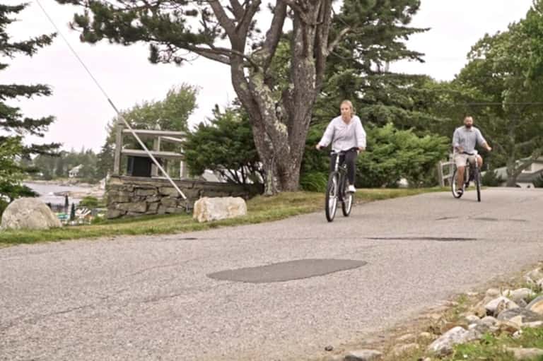 Two people cycling on a residential street near trees and stone structures in a quiet neighborhood