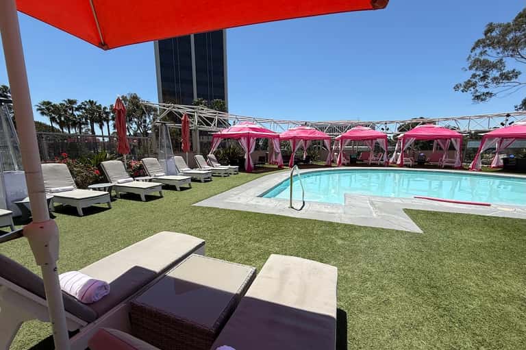 Resort pool area with pink cabanas, green lawn, white loungers, pink umbrellas, and modern building