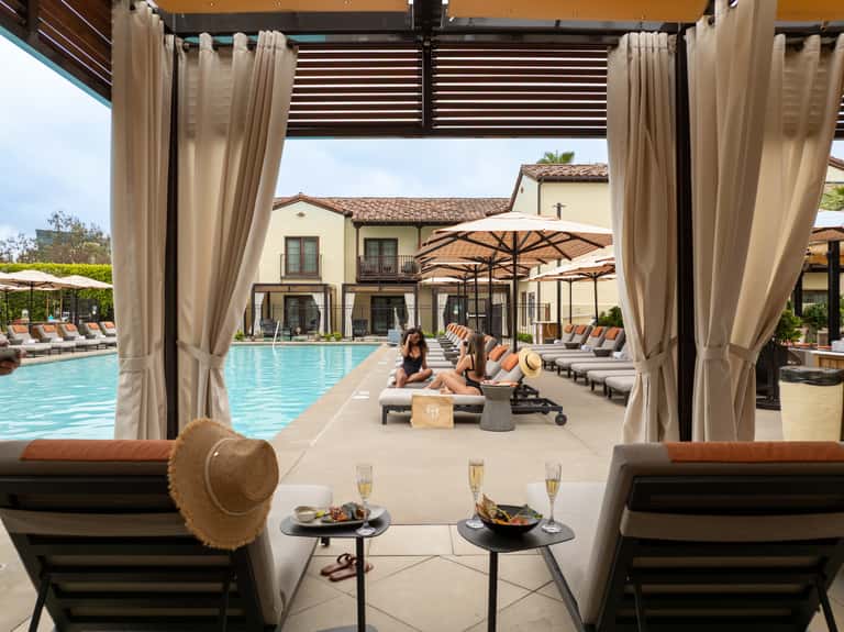 Luxury pool pavilion with cream curtains, lounge chairs, white umbrellas, and Spanish-style architecture visible