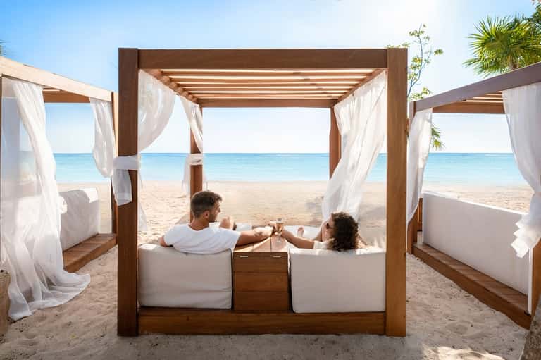 Couple relaxing in wooden cabana bed with flowing white curtains on sandy beach overlooking ocean