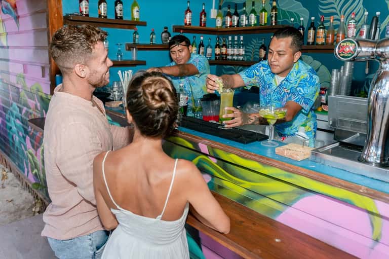 Colorful beachfront bar with turquoise backdrop, bartenders mixing drinks for guests