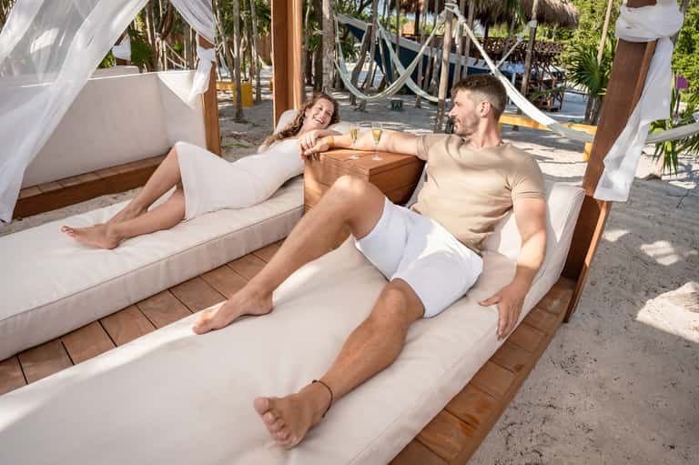 Couple relaxing in beachfront cabana with white curtains and ocean view at sunset
