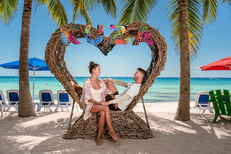 Couple posing at heart-shaped swing on tropical beach with umbrellas and palm trees