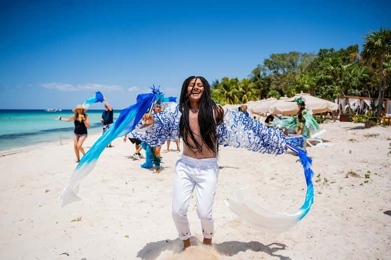 Woman in blue and white outfit dancing on tropical beach with turquoise ocean and guests