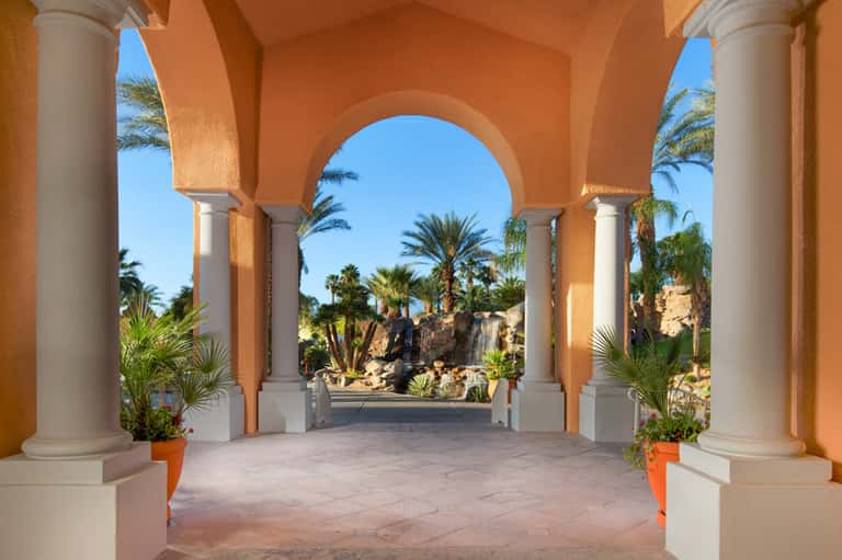 Orange colonnade with arched ceiling overlooking palm trees and rocky landscape
