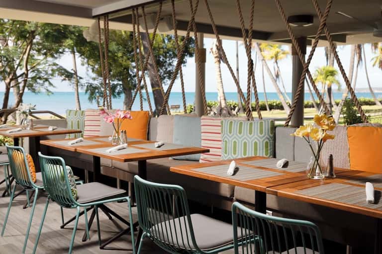 Beachfront dining area with rope swings, ocean view, colorful cushions, and wooden tables with place settings