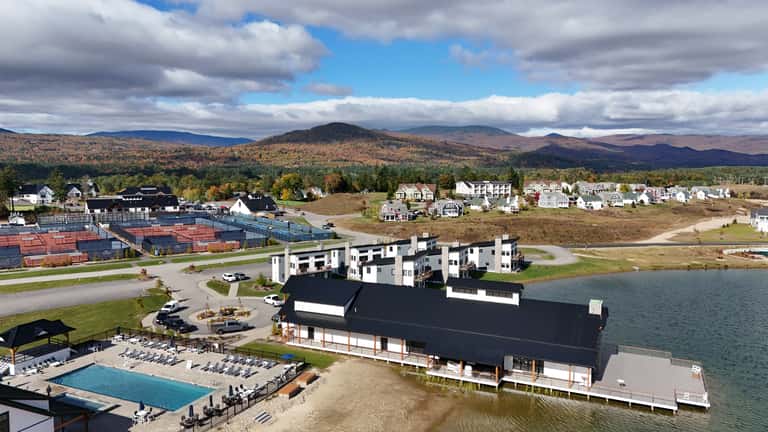 Aerial view of lakeside resort with pool, white hotel building, mountains, forests, and sports courts