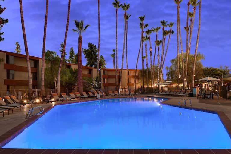 Illuminated resort pool at dusk with palm trees, multi-story building, lounge chairs, and glowing water