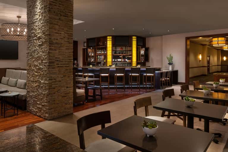 Upscale resort bar and lounge area with brick column, dark wood furnishings, and illuminated bar counter
