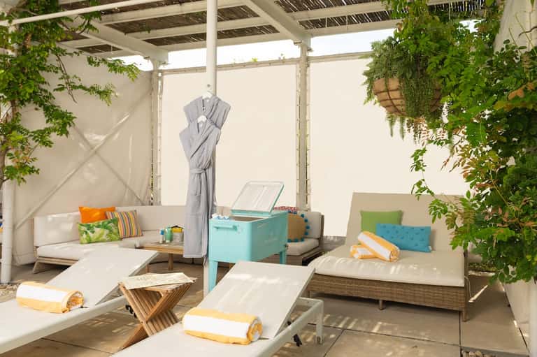 Covered outdoor lounge with daybed seating, colorful pillows, cooler, and hanging umbrella