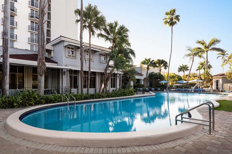 Resort pool with curved edge, palm trees, white building, and sunny afternoon setting