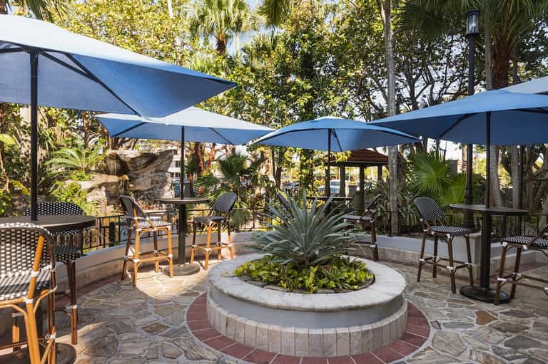 Tropical outdoor patio with palm trees, blue shade sails, fire pit, and casual seating