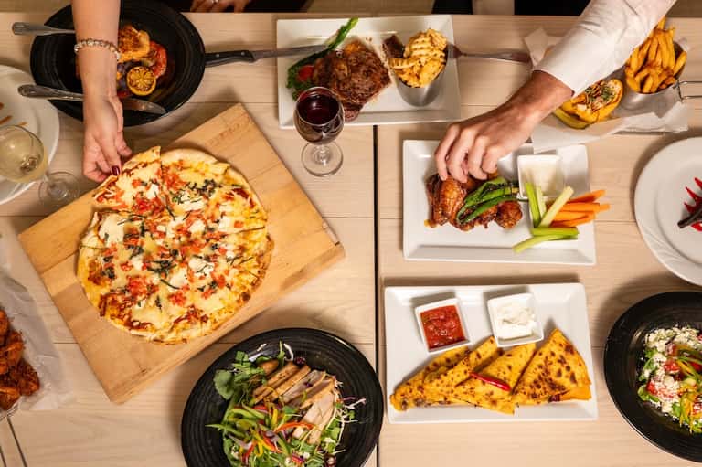 Overhead view of restaurant table with pizza, grilled steak, fries, and wine glasses shared by diners